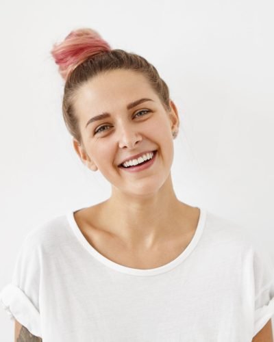 Cheerful female model with hair bun colored in pink, wearing casual clothes, smiling broadly while being happy to recieve gift. Attractive female having good mood isolated over white background